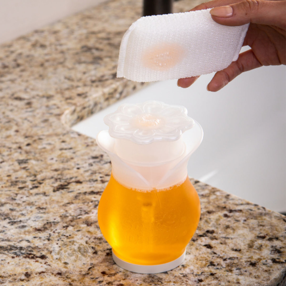 Orange drink in a glass with a white lid being wiped by a hand holding a white cloth on a marble countertop.
