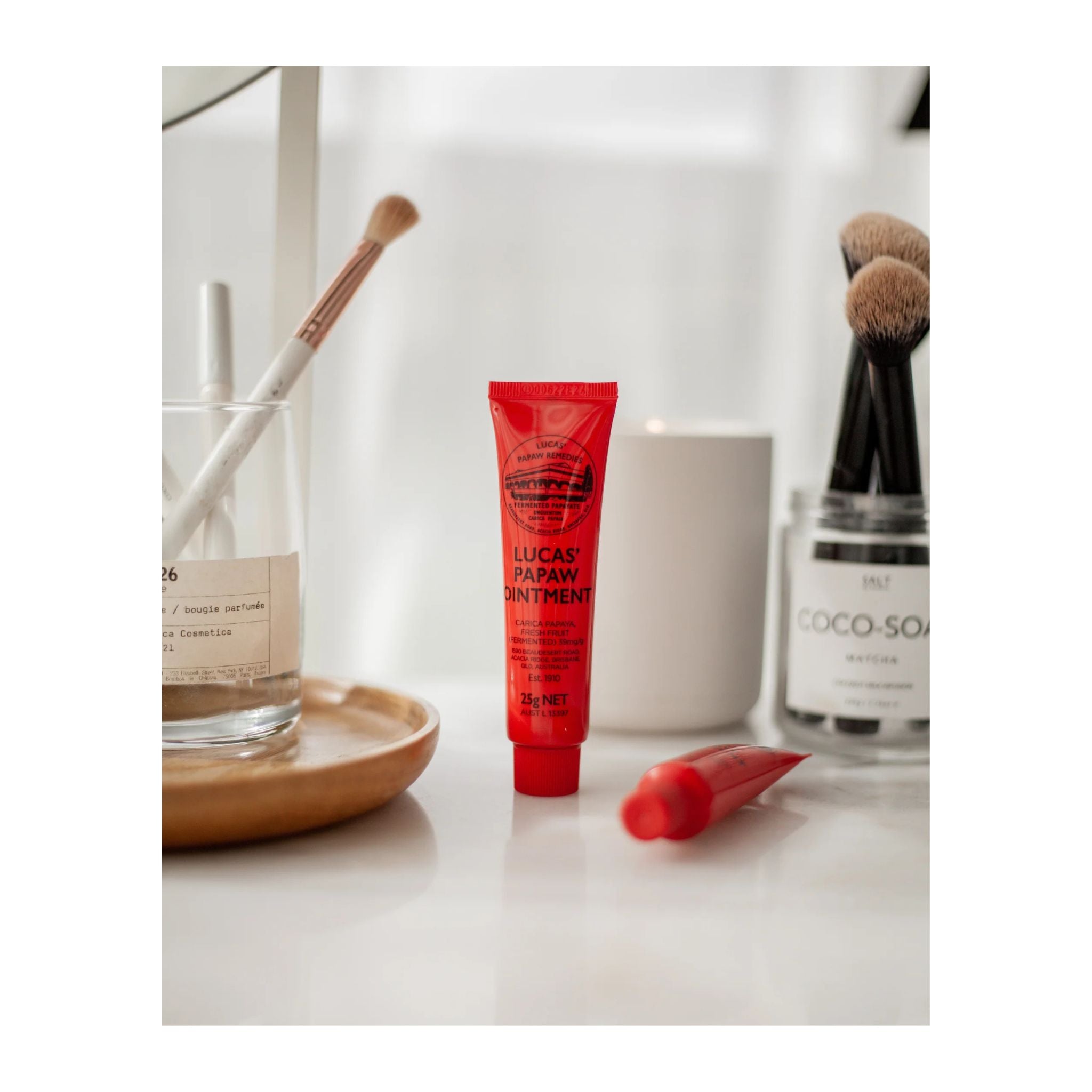 Red tube of Lucas Papaw Ointment on a white surface with makeup items in the background