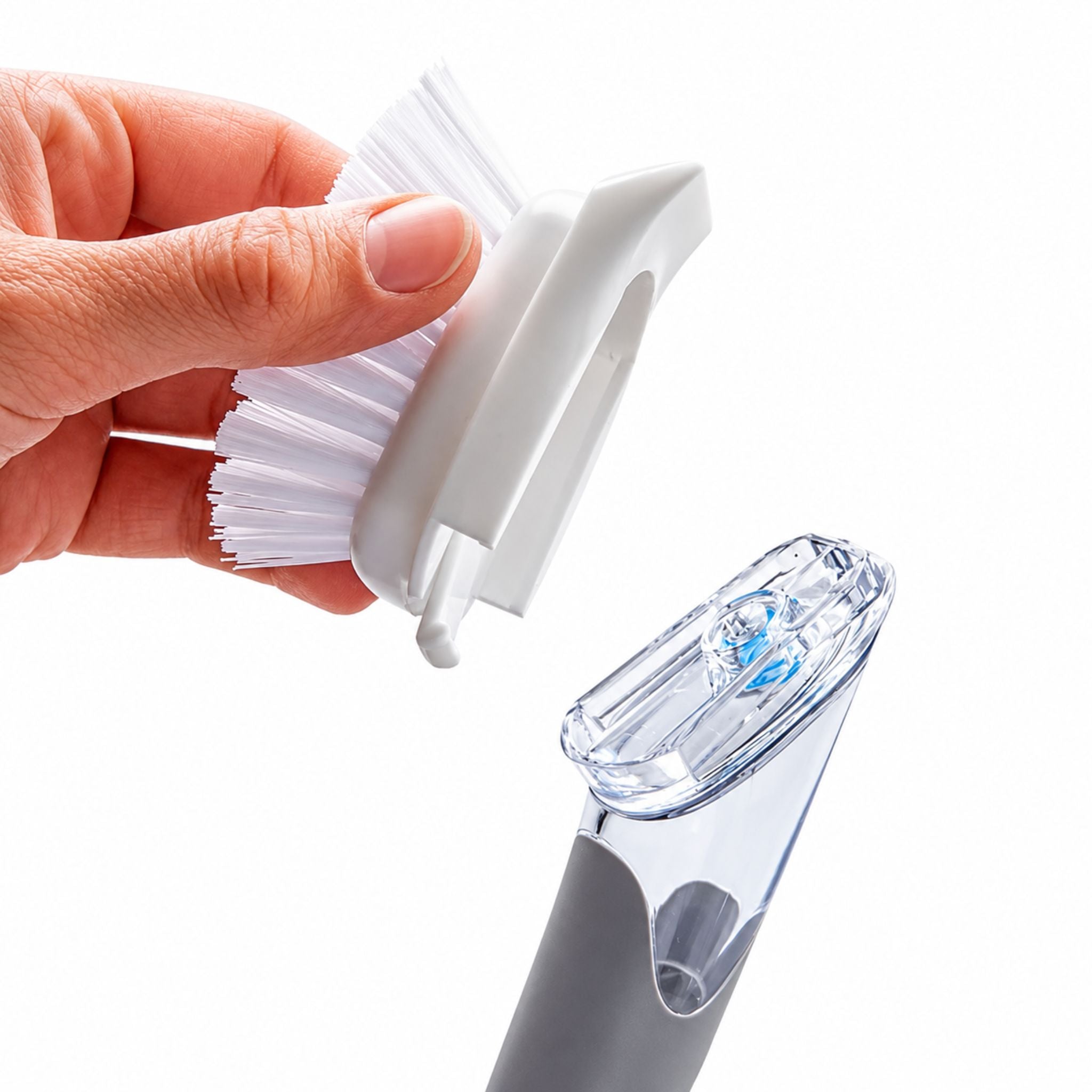 A hand is shown holding a white plastic brush head with white bristles, demonstrating how it attaches to a transparent and gray handheld cleaning device on a white background.